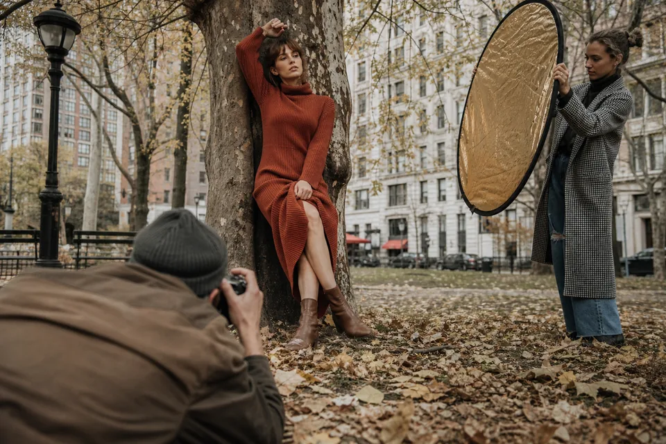 Plener w parku jesienią, fotograf z aparatem i asystentka ze złotą blendą przed dorosłą modelką